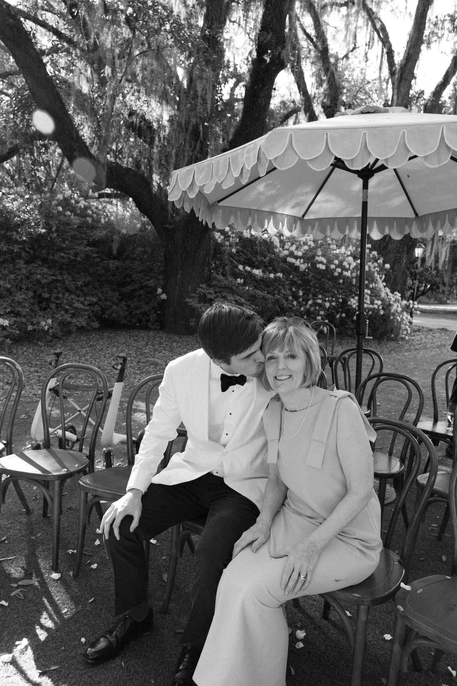 A couple sits together on garden chairs beneath a white decorative umbrella in a black and white outdoor setting.