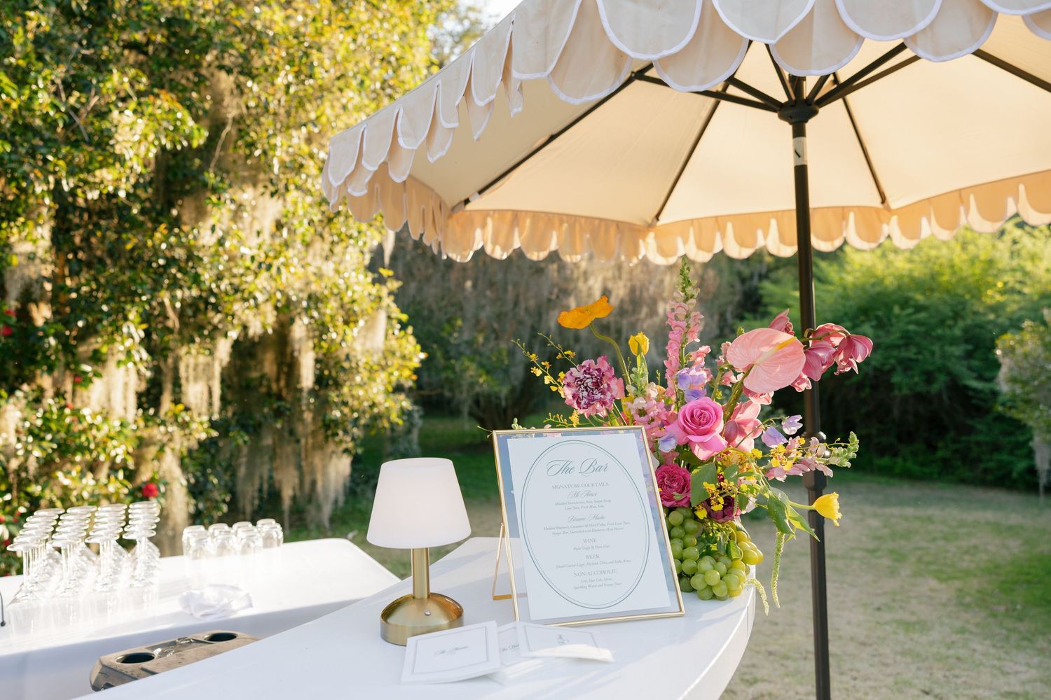 Outdoor party setup with white umbrella, table lamp, and colorful floral arrangement under sunlit trees.