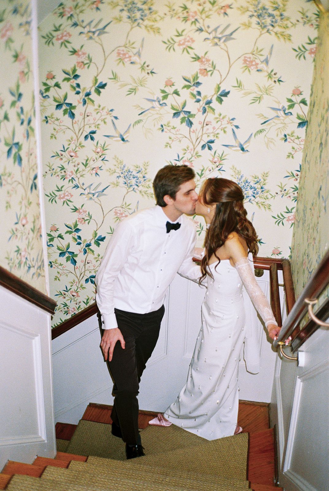 A couple shares a romantic kiss on a staircase with floral wallpaper featuring delicate blue and green botanical patterns.
