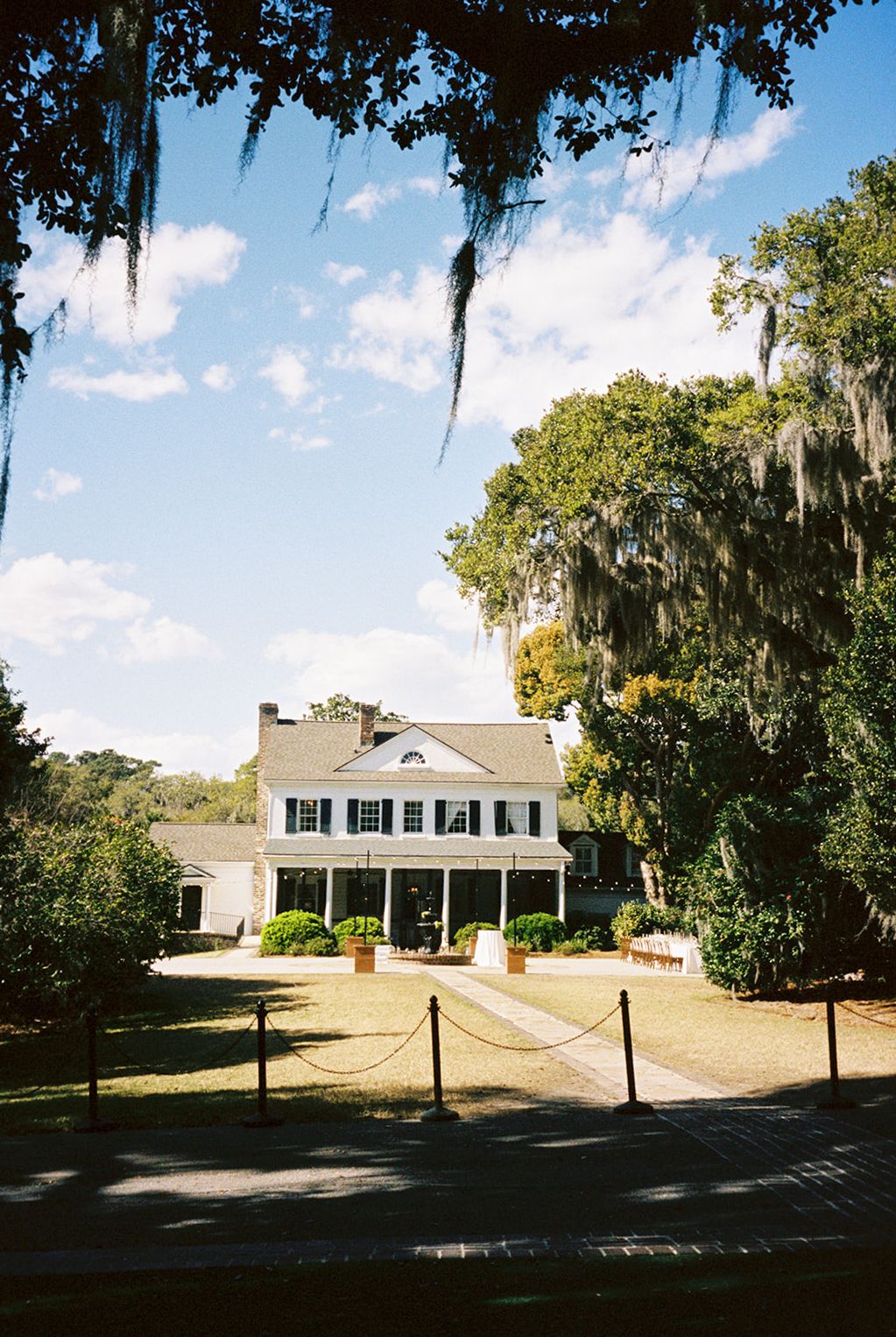 A stately white Southern plantation house with Spanish moss draped oak trees and a roped entrance path on a sunny day.