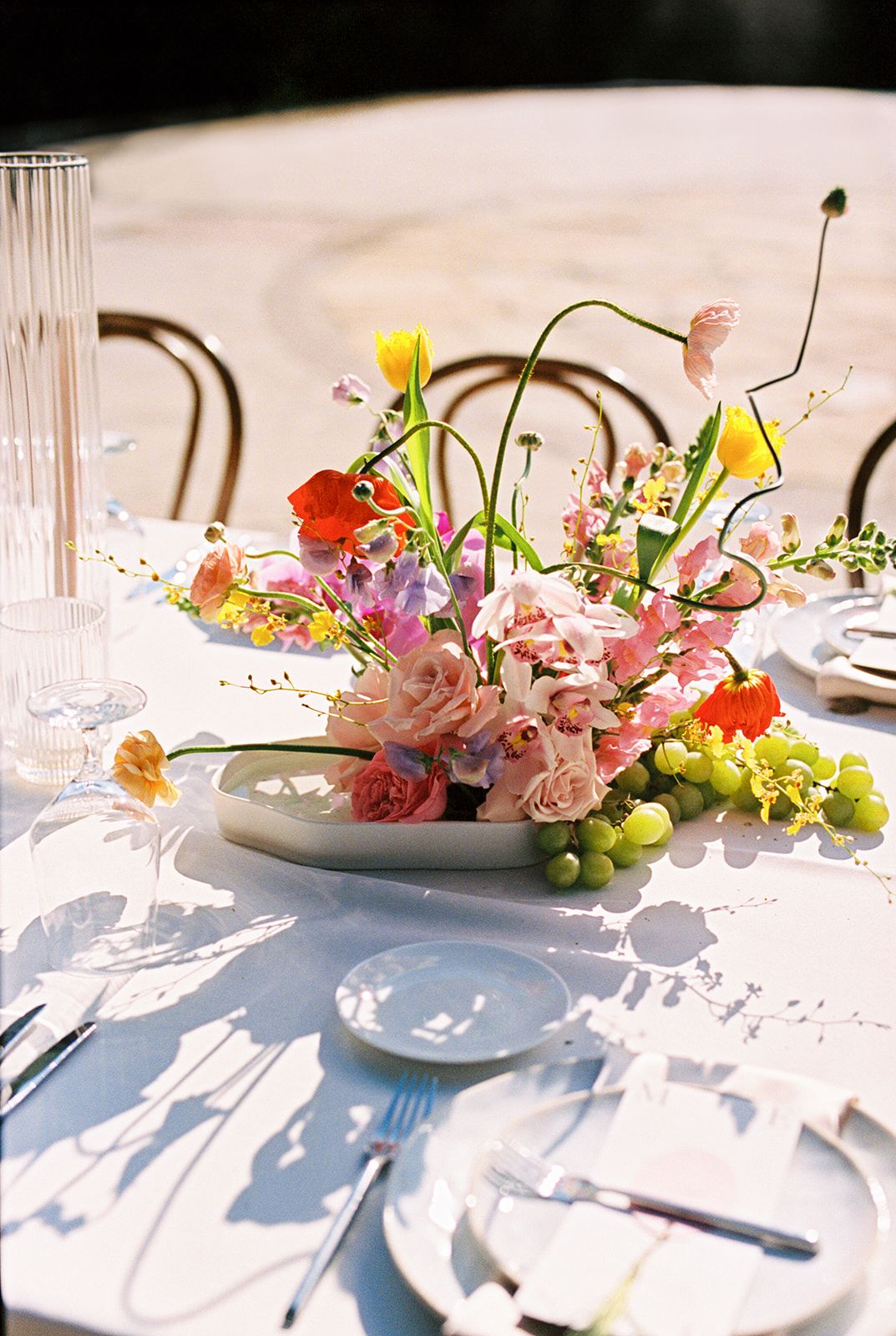 An elegant table setting with a floral centerpiece of pink and yellow flowers, grapes, and crystal glassware in natural light.