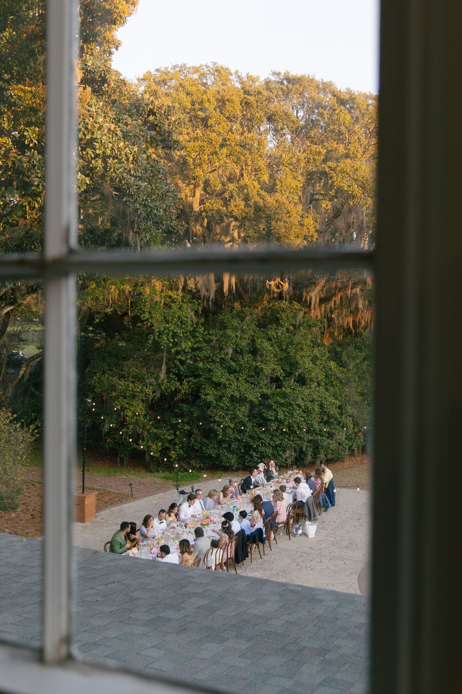 A view through a window showing a large group dining at a long table outdoors during golden hour.