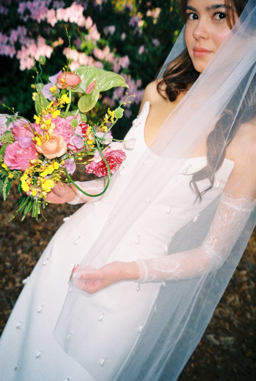 A bride in a white gown holding a vibrant pink and yellow floral bouquet with flowing tulle veil in an outdoor setting.