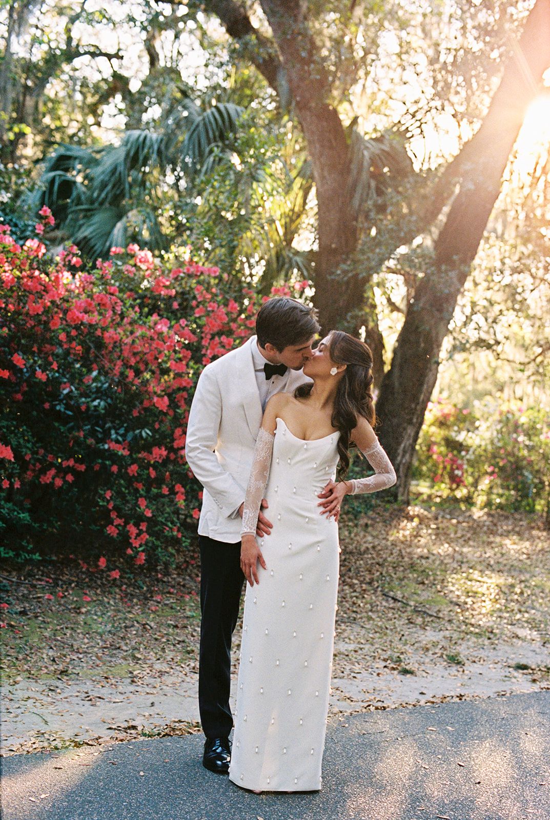 A romantic wedding photo captured in a garden setting with blooming red flowers and sunlight filtering through trees.