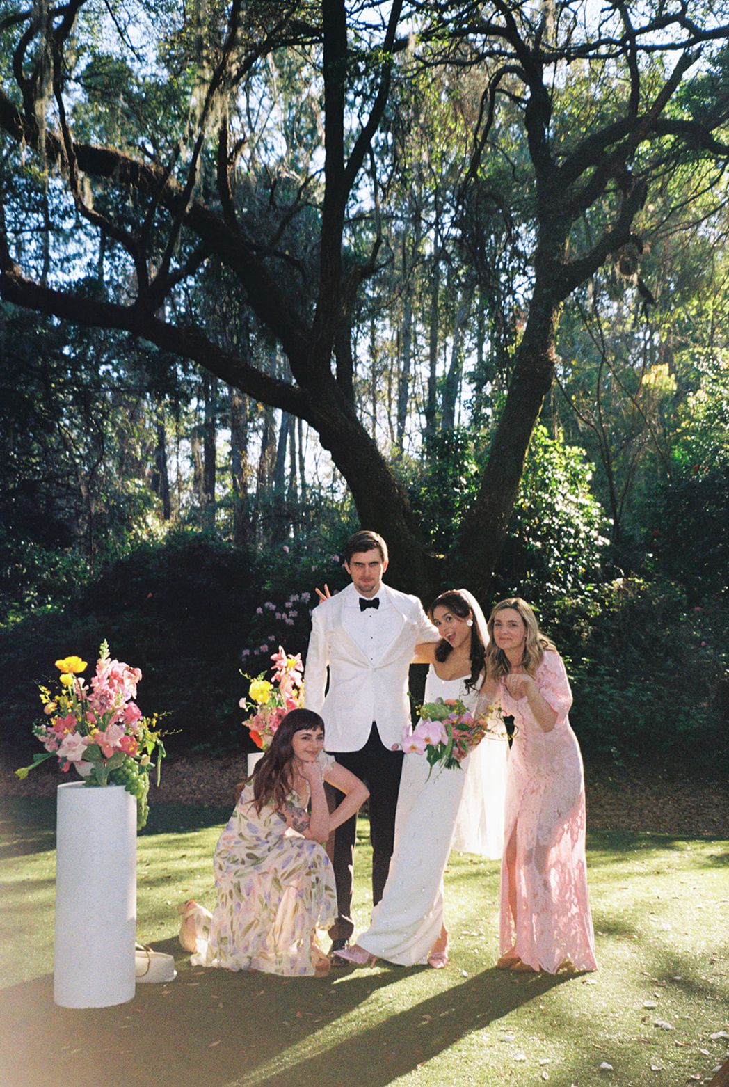 A group poses together at an outdoor garden wedding ceremony with floral arrangements and tall trees in the background.