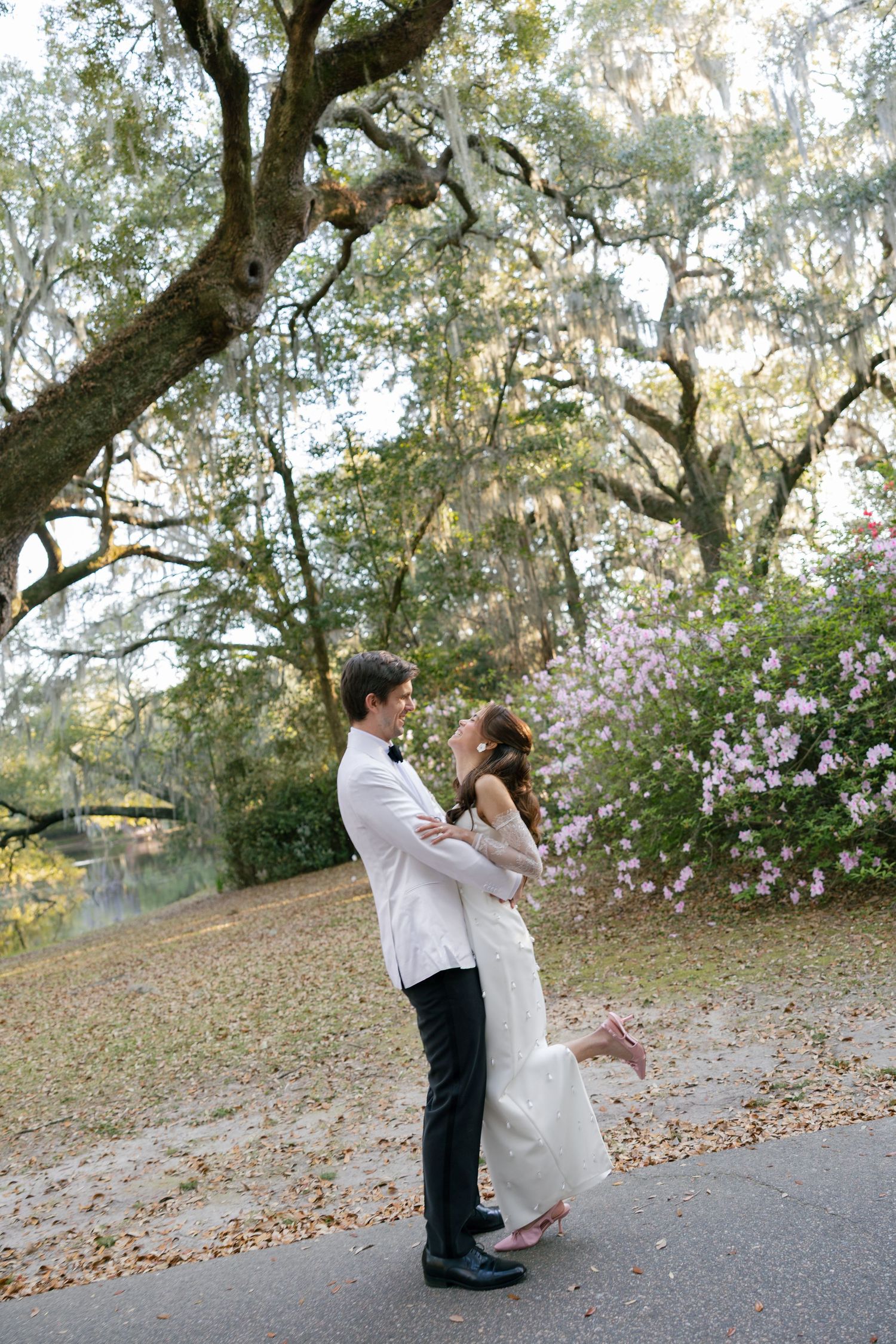 A romantic couple embraces on a garden path under flowering trees and Spanish moss in a scenic outdoor setting.