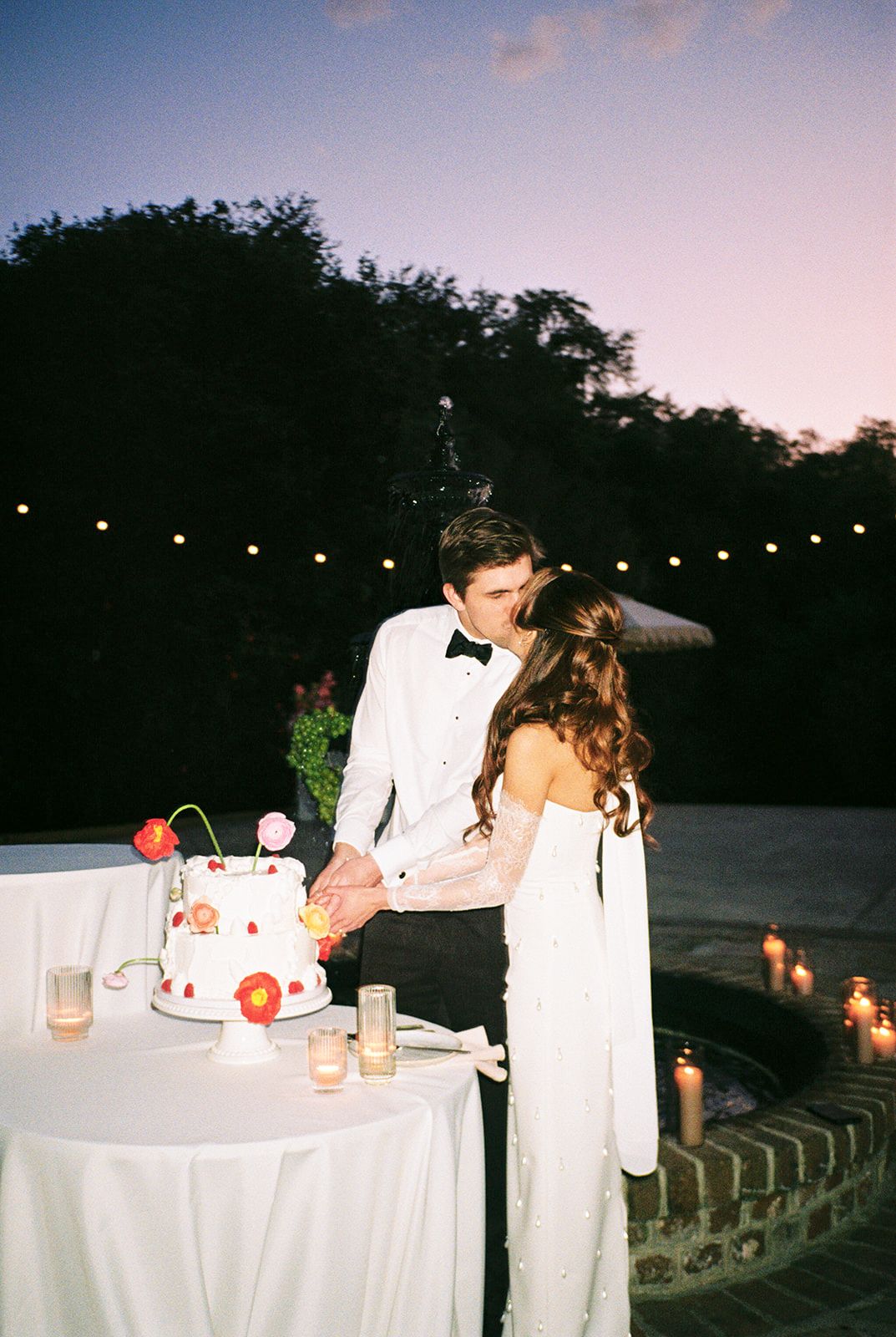 A couple cuts their wedding cake together at an outdoor evening reception under string lights at dusk.