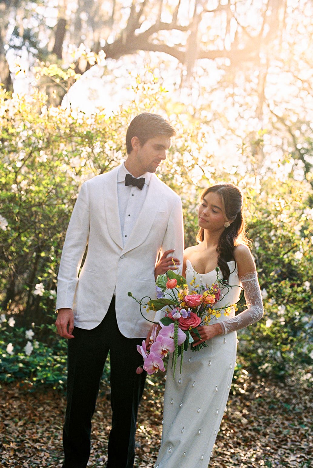 Elegant couple in formal wedding attire stands in a sunlit garden with a colorful bouquet of flowers.