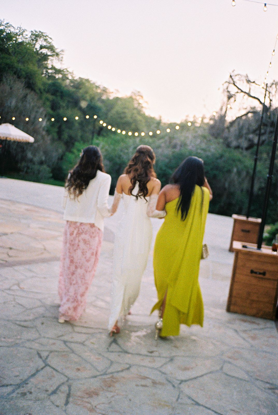 Three women in flowing dresses walk together on a stone patio with string lights overhead at dusk.