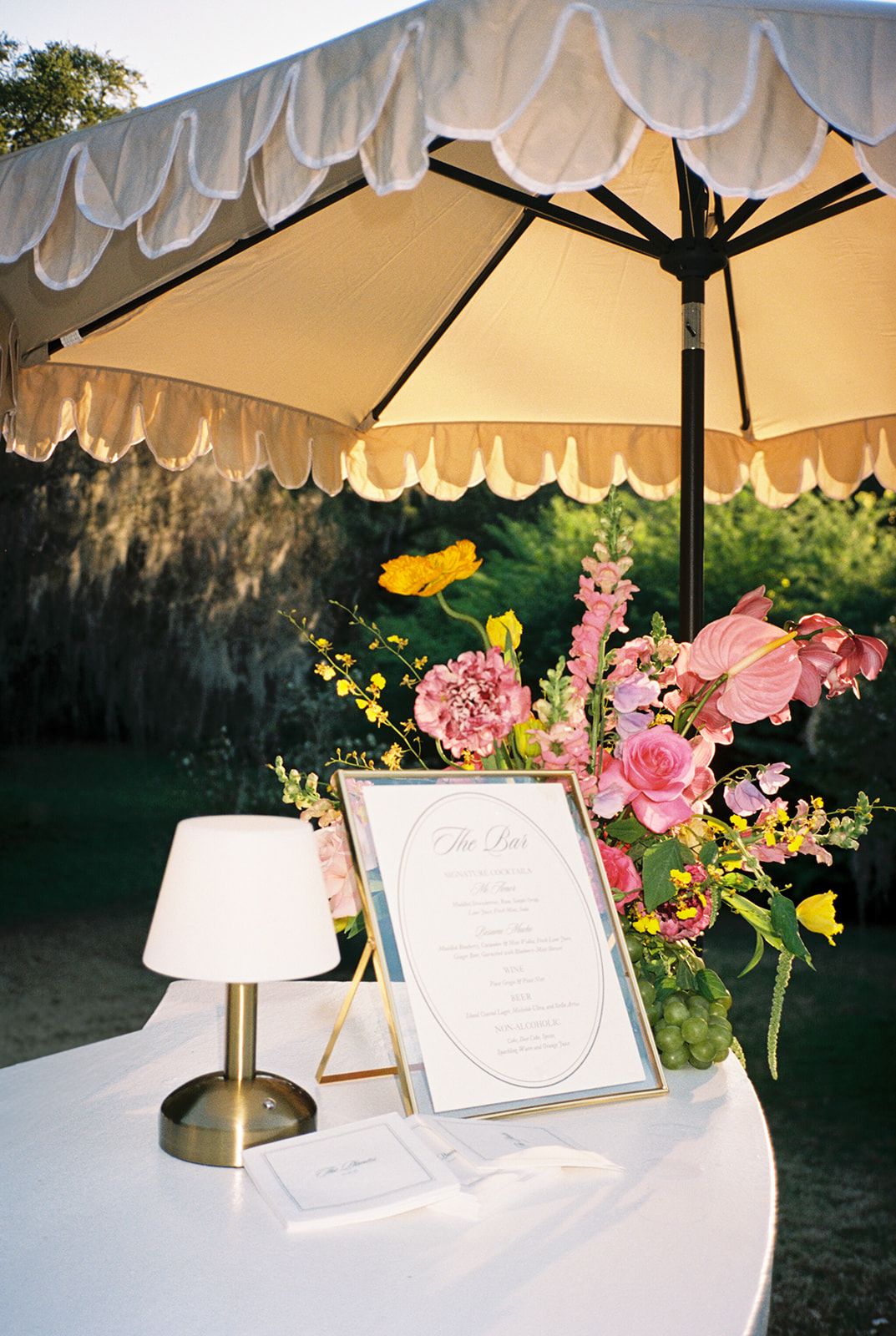 Elegant outdoor table settings with pink and yellow floral arrangements under white umbrellas for an upscale garden event.