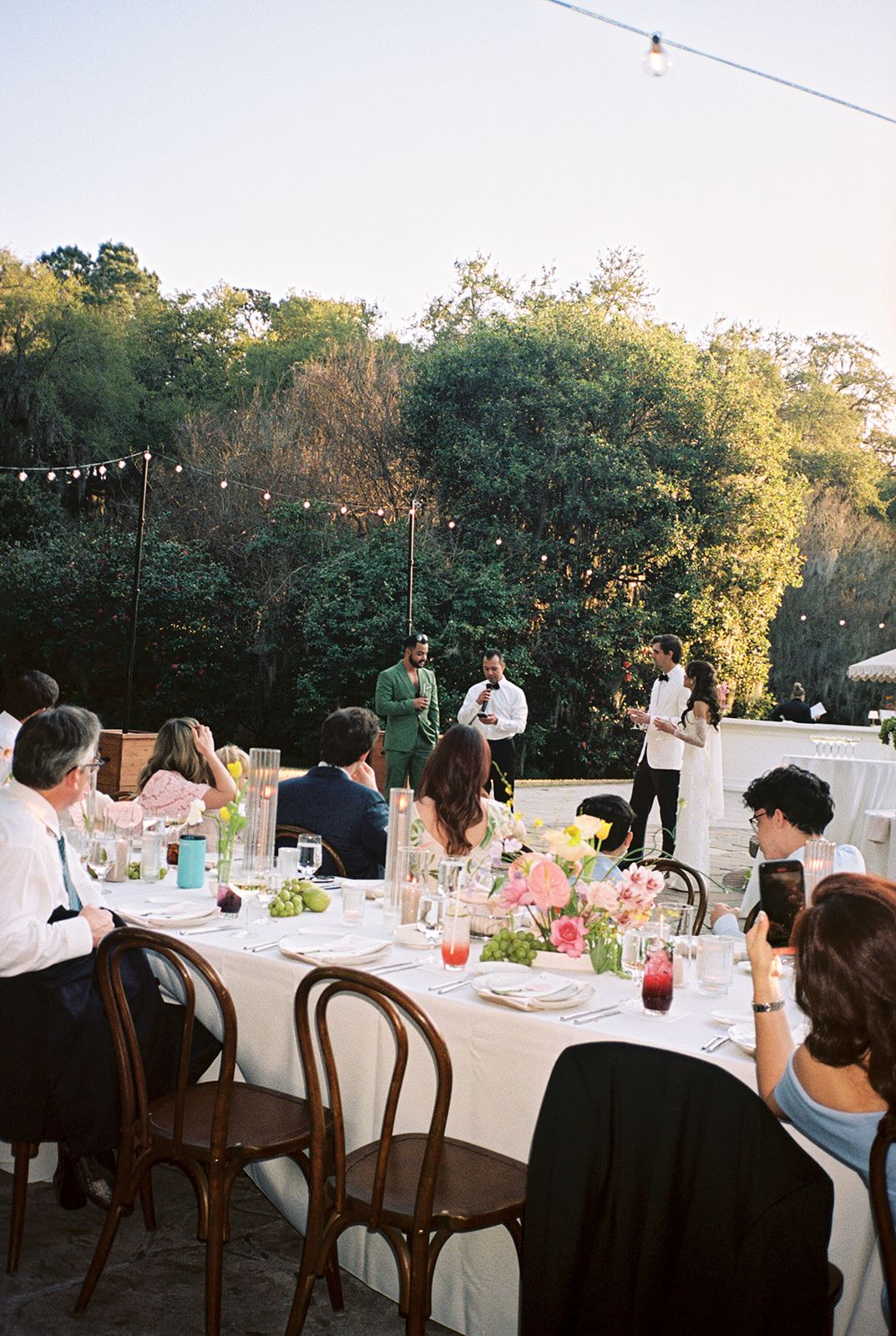 Outdoor wedding reception tables decorated with white linens and floral centerpieces under string lights at dusk.
