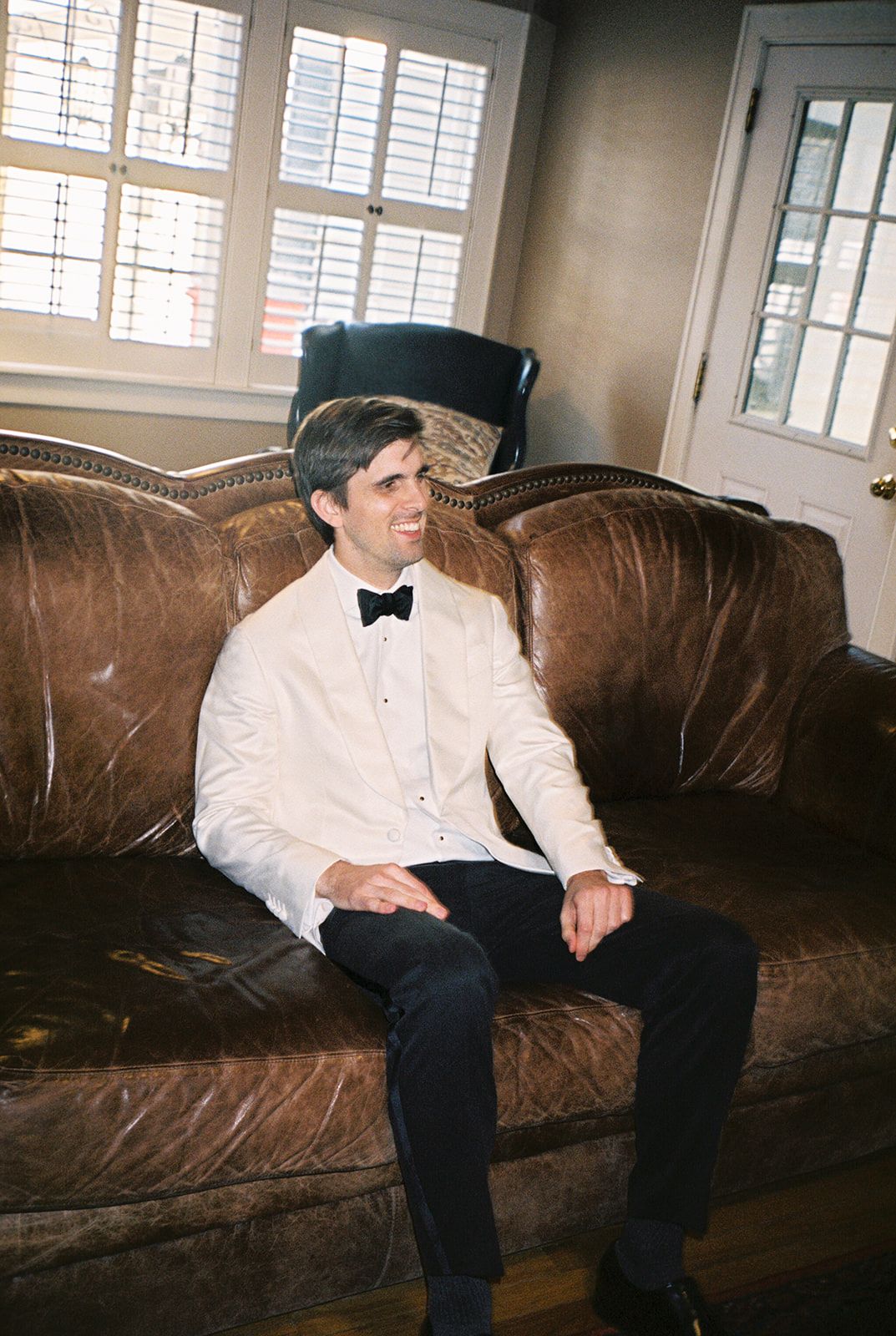 A person in formal attire with a white dinner jacket and bow tie sits relaxed on a brown leather couch in a bright room.