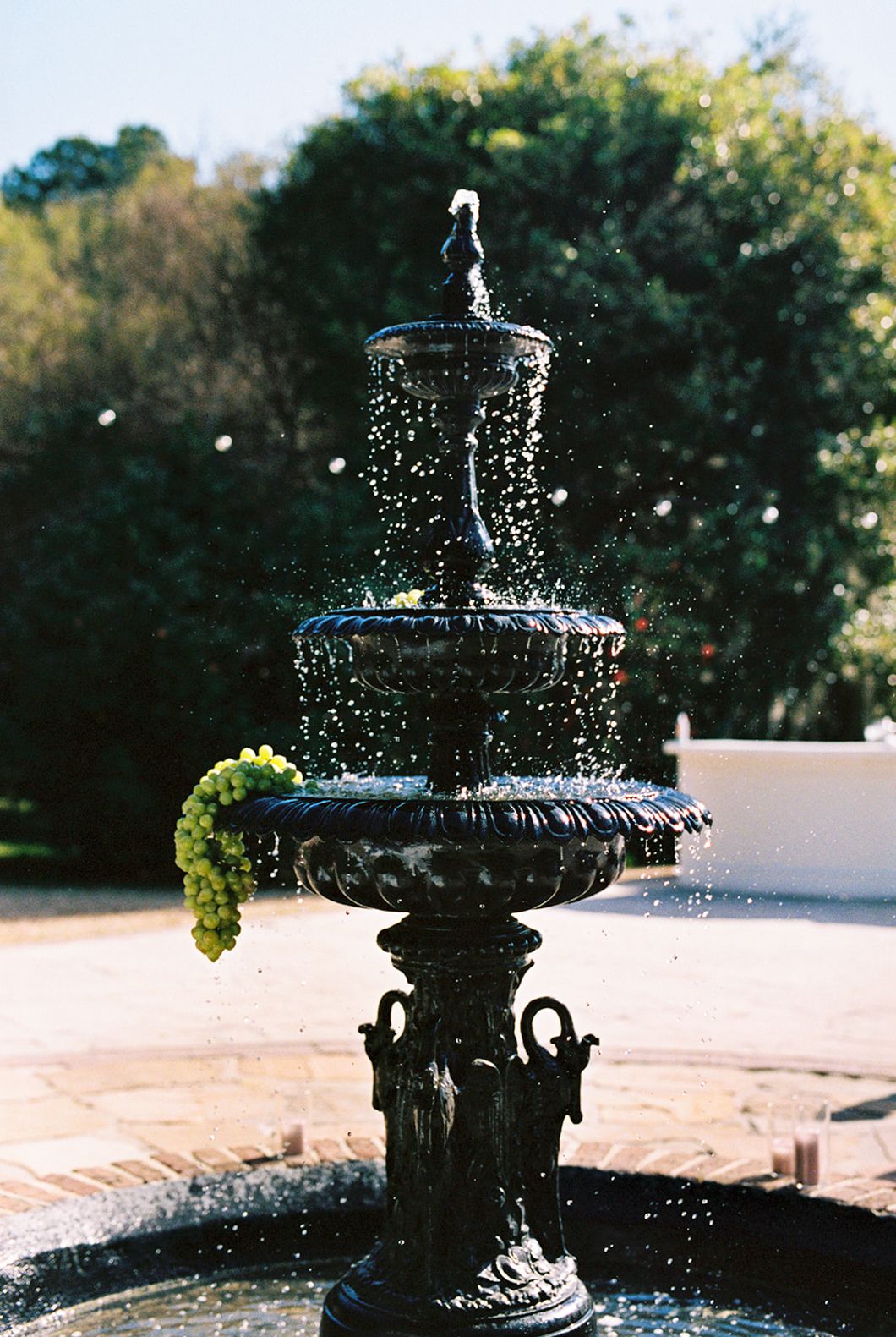 Ornate black tiered fountain spraying water with sunlit droplets against green foliage background in garden setting.