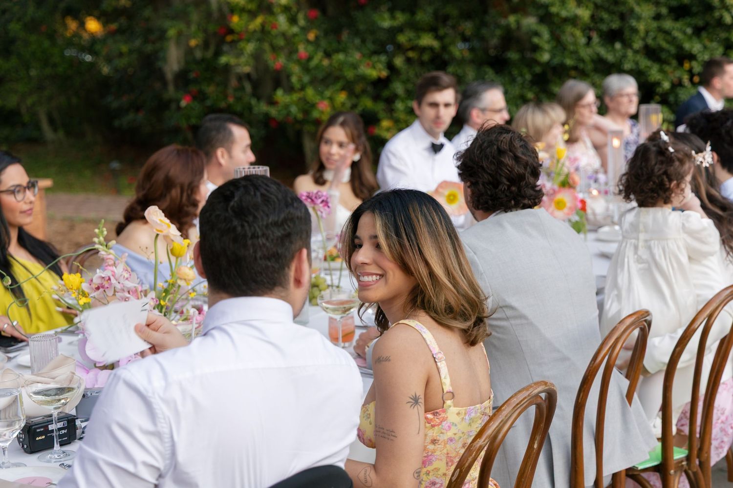 Wedding guests sitting at a long outdoor dinner table decorated with floral arrangements during a garden reception celebration.