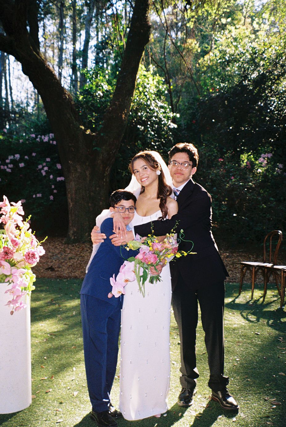 A small outdoor wedding ceremony in a wooded garden setting with floral arrangements and gentle sunlight.