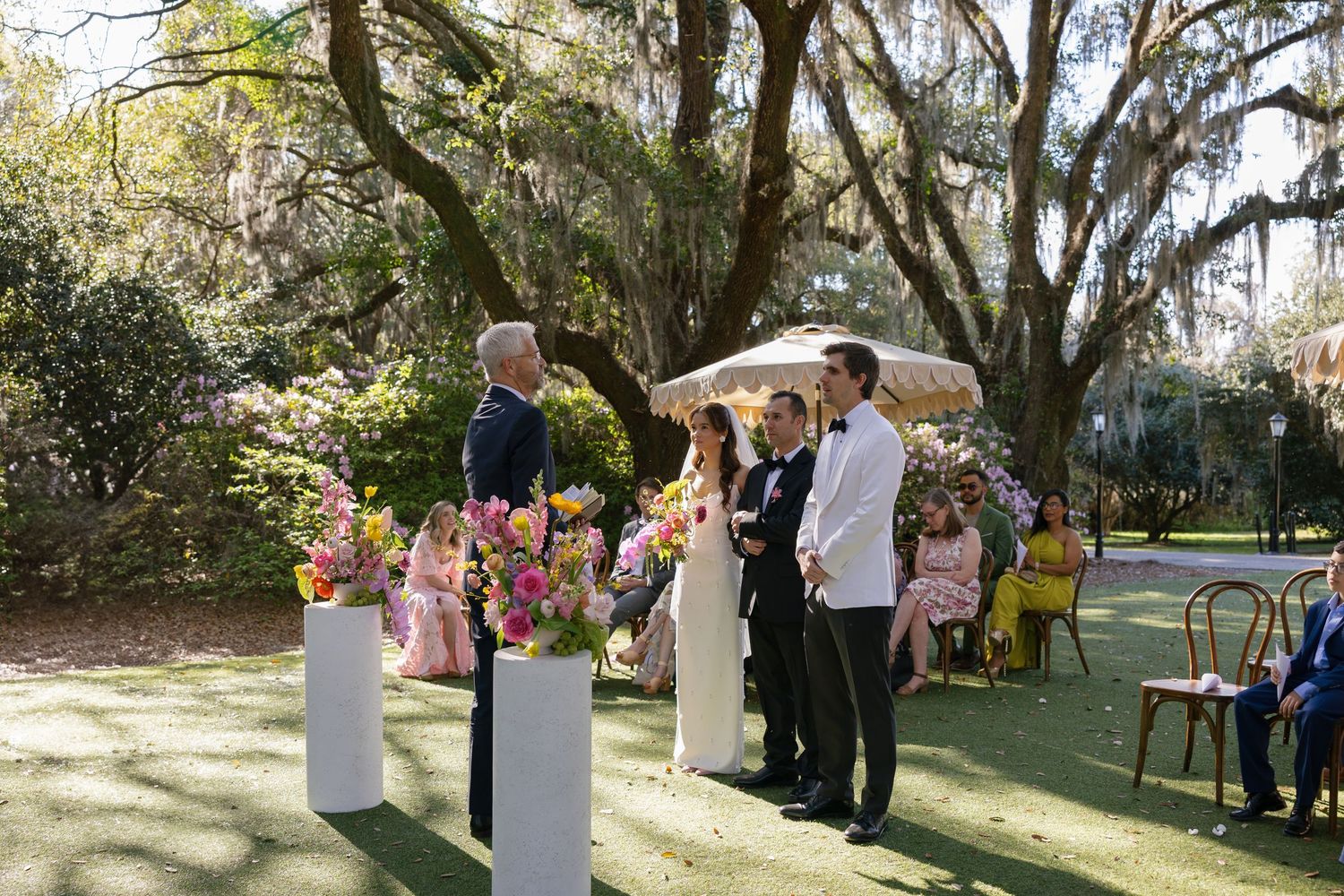 A scenic outdoor wedding ceremony takes place under large oak trees with guests gathered to witness the occasion.