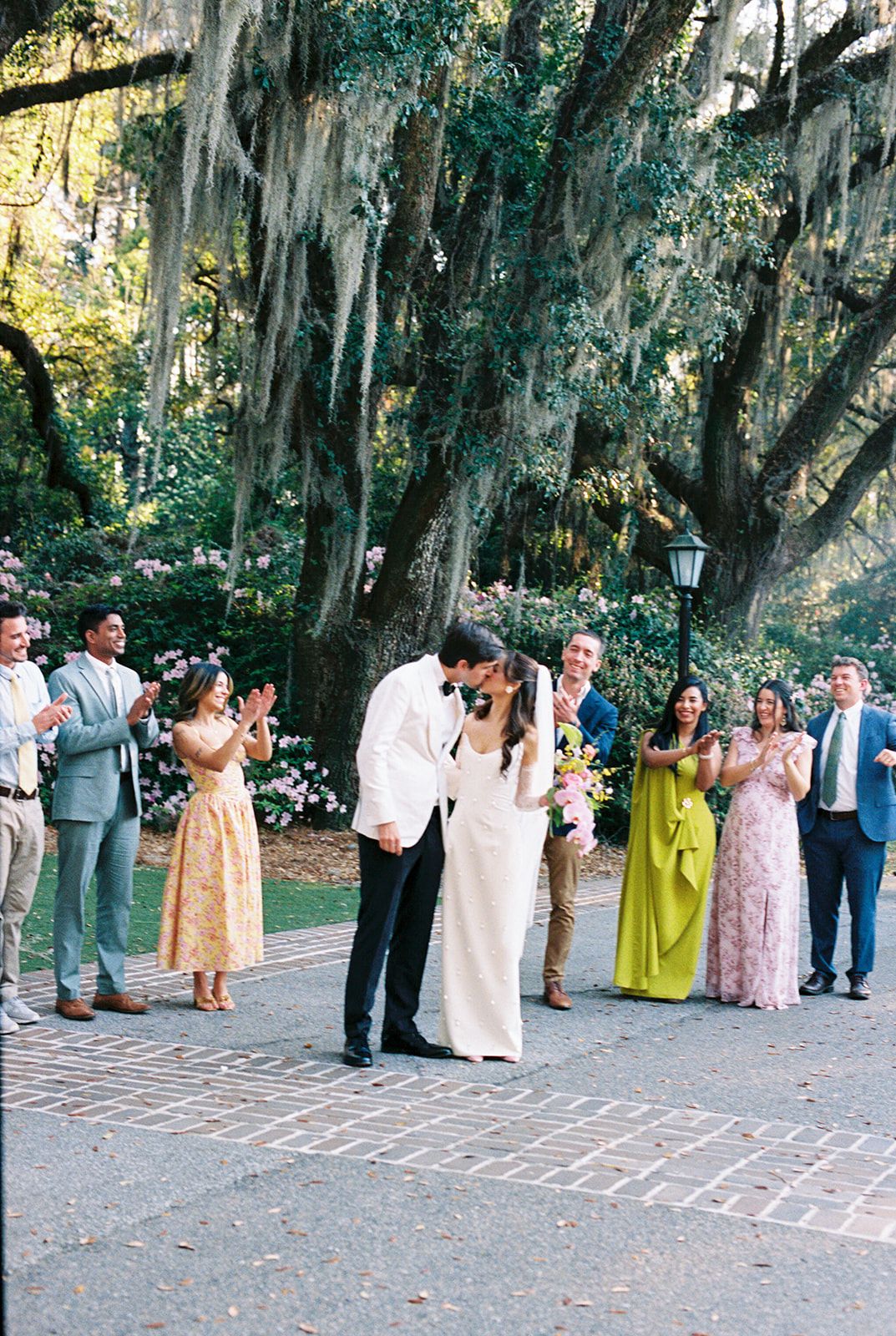 Wedding guests gather in a line beneath Spanish moss-draped trees during an outdoor ceremony in a garden setting.