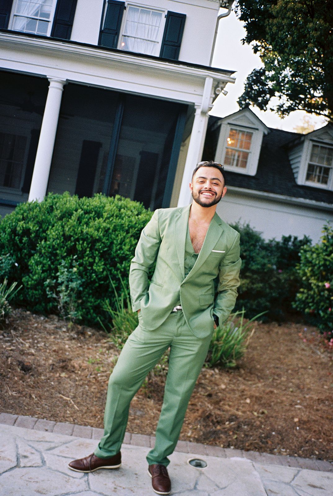 A person in a mint green suit strikes playful poses in front of a white house with a garden.