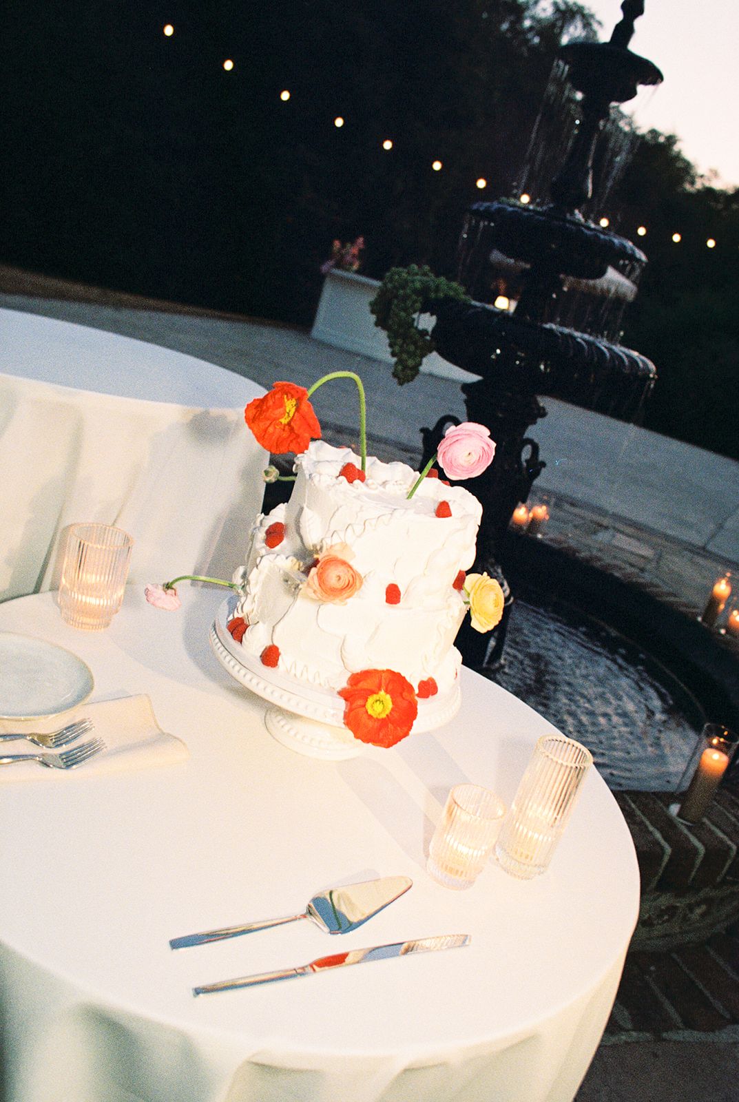 A white tiered wedding cake decorated with orange and pink poppies sits on a round table at an outdoor evening reception.