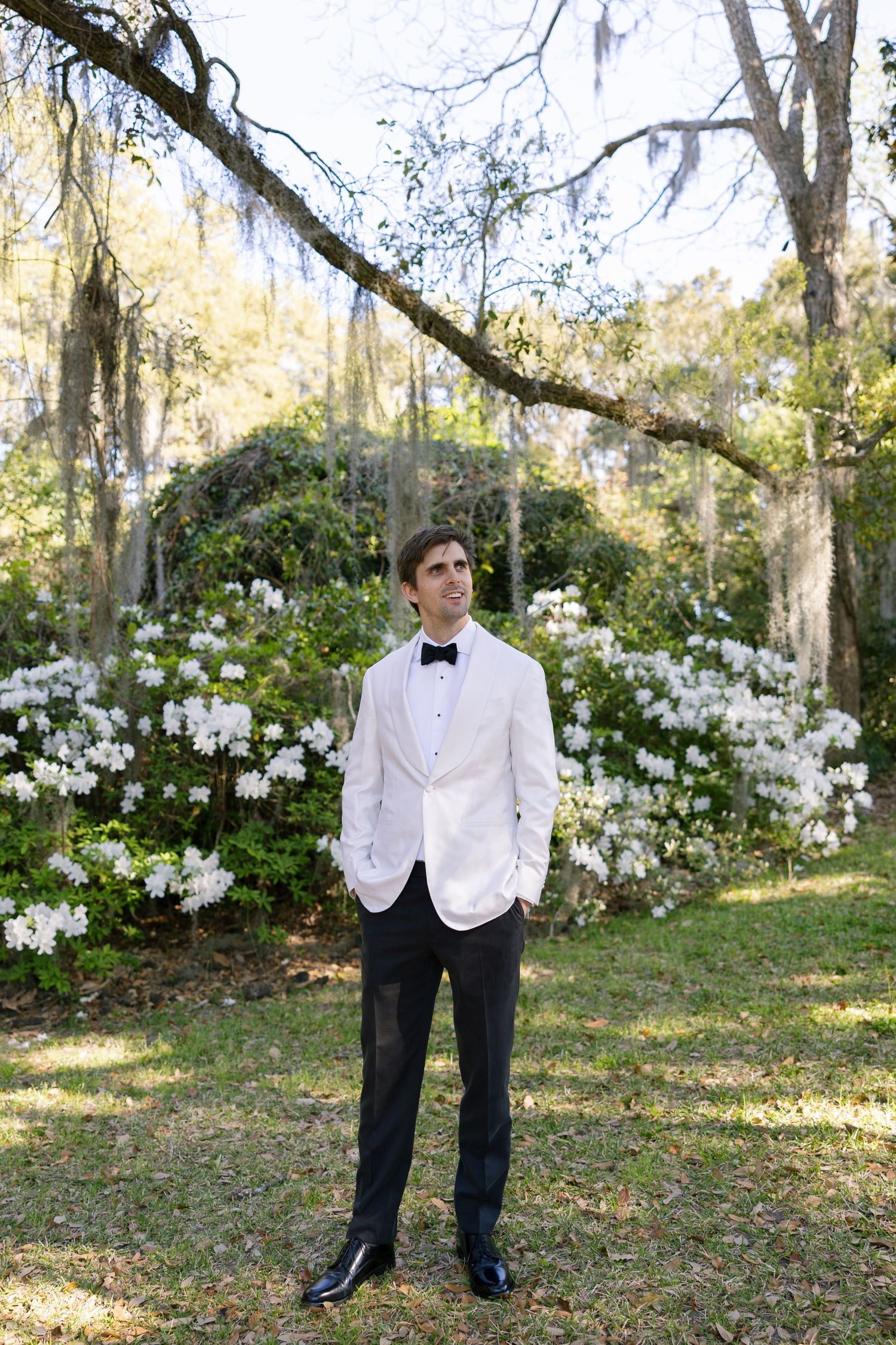 Someone in a formal white dinner jacket and bow tie standing in a garden with blooming white azalea bushes and Spanish moss trees.