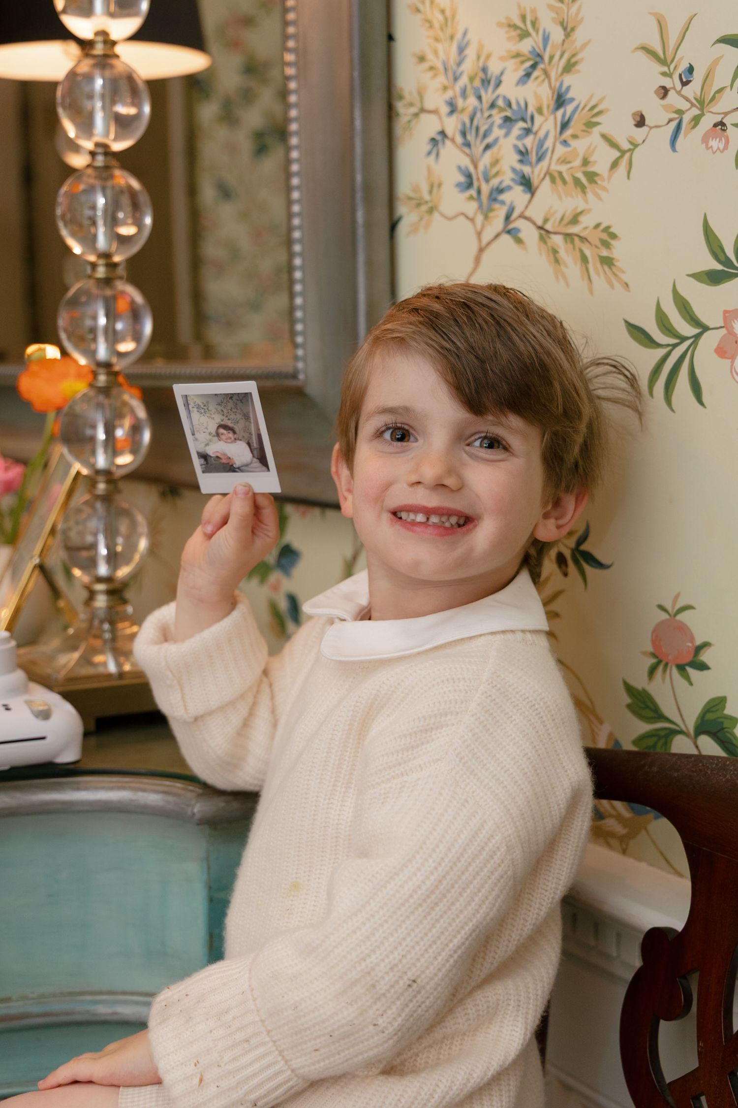 Young child in cream sweater sits at desk with floral wallpaper and glass lamp, holding a small photograph.