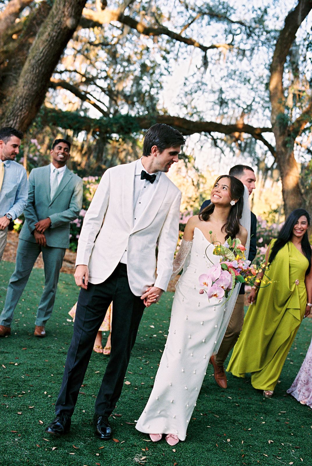 A joyful wedding party dances and celebrates together on a grassy lawn surrounded by trees during golden hour.