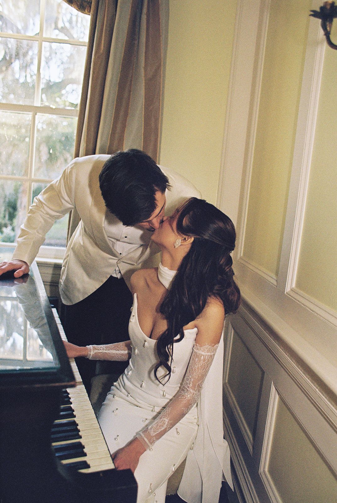 A romantic couple in wedding attire share a kiss while sitting at a piano in a softly lit room.