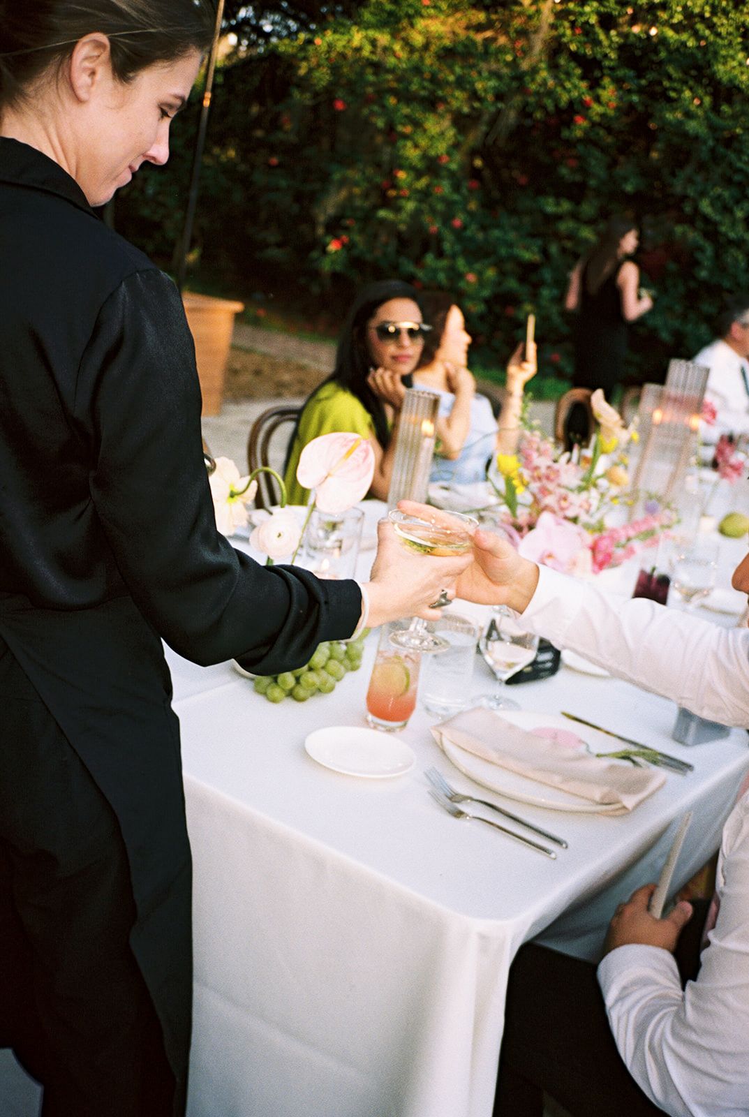 Waiter in black uniform serving guests at elegant outdoor table setting with crisp white linens under tree canopy.