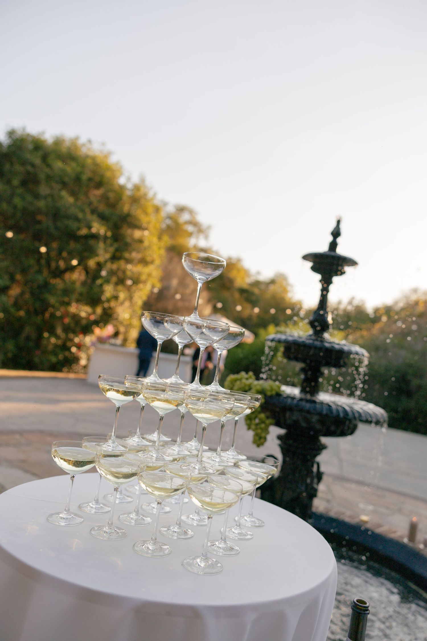 Pyramid of elegant champagne glasses stacked on white tablecloth next to ornate black fountain in outdoor garden setting.