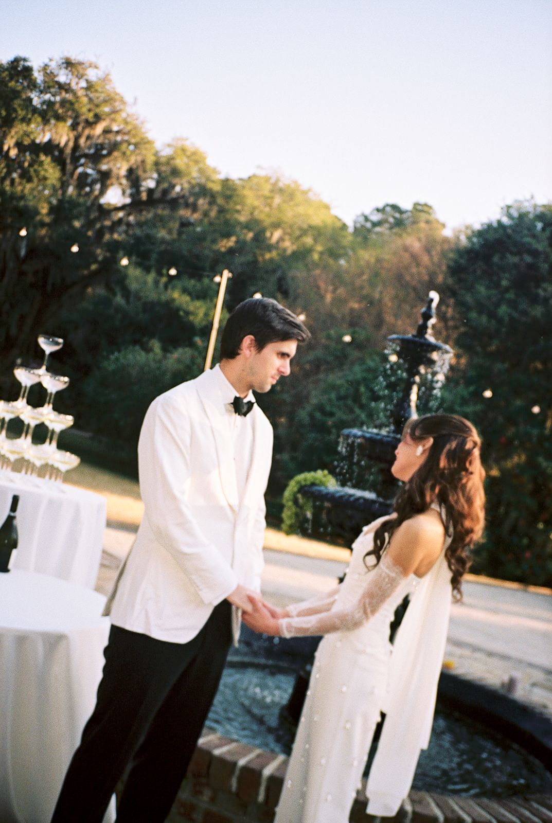 A couple in formal wedding attire dances and embraces in a romantic outdoor garden setting at sunset.