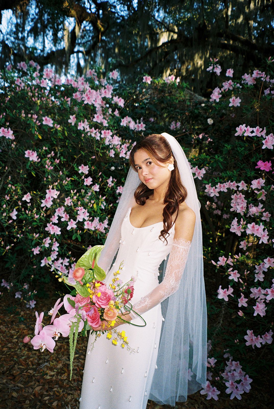 A bride in a white wedding dress poses elegantly among blooming pink azalea flowers in a garden setting.