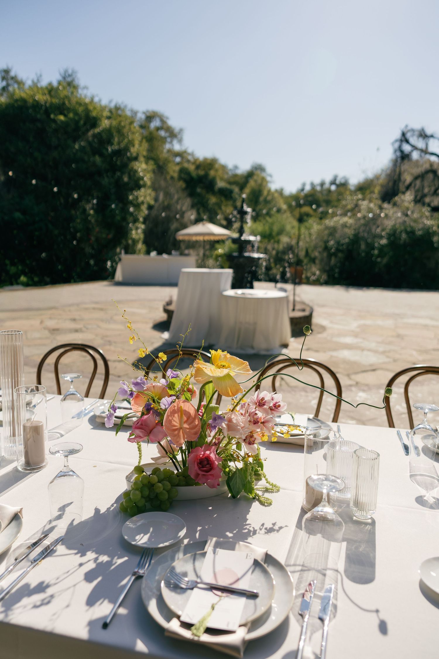 An elegant outdoor table setting with a colorful floral centerpiece and bentwood chairs under string lights and trees.