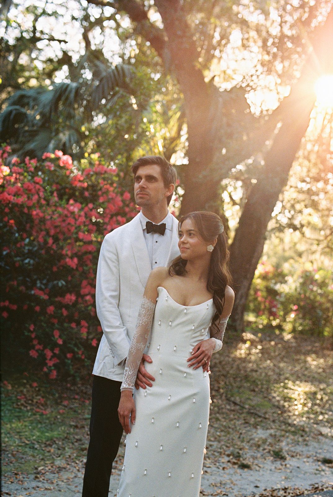 A romantic wedding photo captured at sunset with pink flowers and trees in the background, displaying elegant attire.