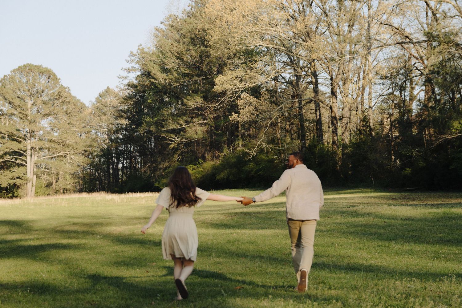 Two people run together holding hands across a sunlit grassy field bordered by tall trees on a warm day.