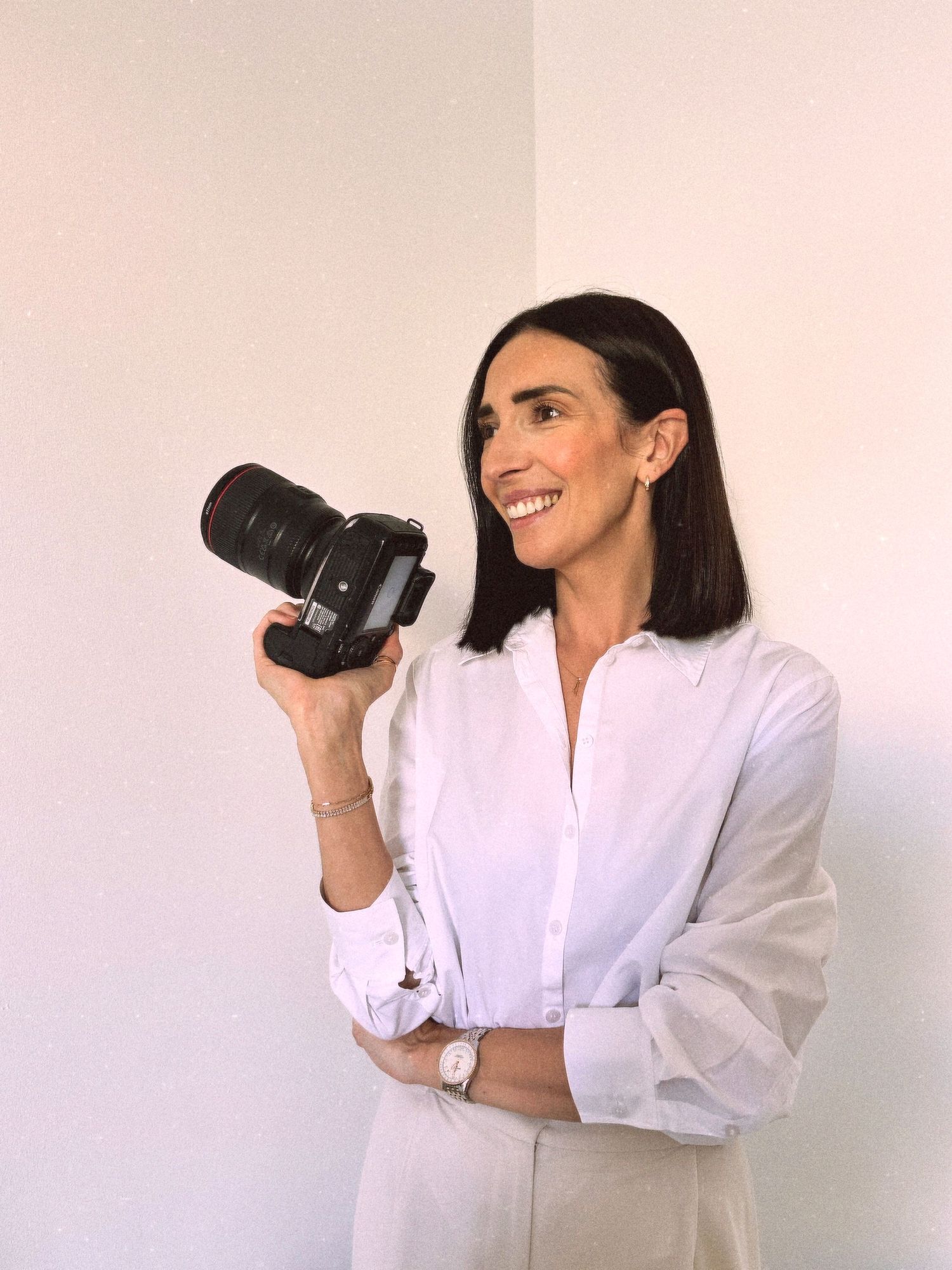 A photographer in a white blouse holds a professional DSLR camera against a pale pink wall.