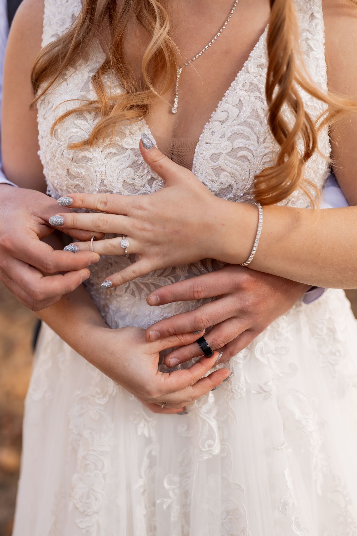 Close-up of hands wearing wedding rings against a detailed lace white wedding dress.