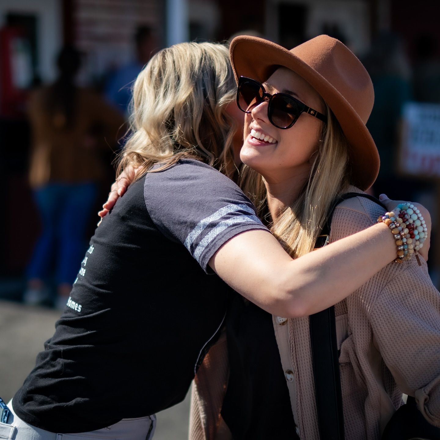 Two friends sharing a warm embrace outdoors, one wearing a brown hat and sunglasses.
