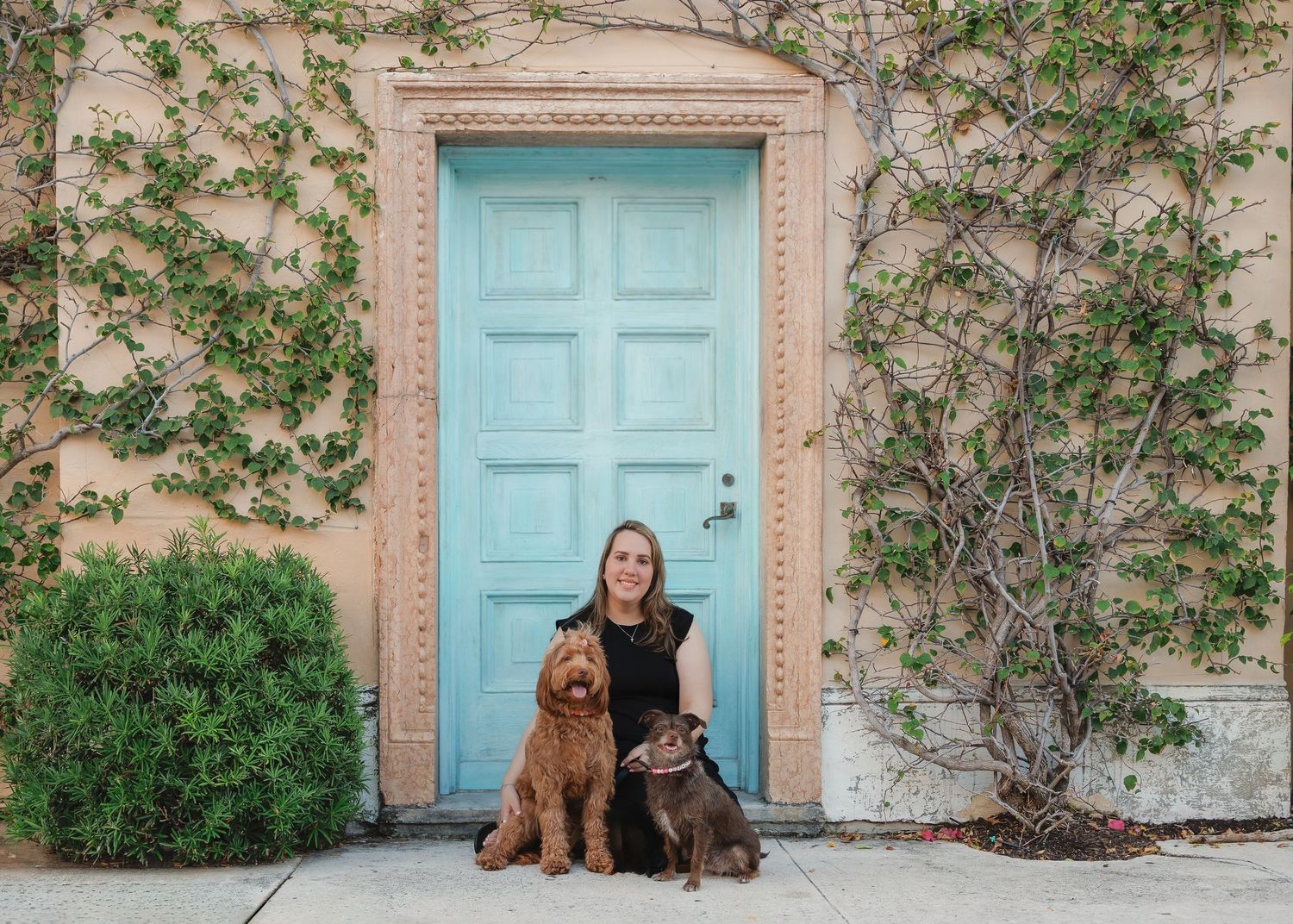 Solo portrait with dogs sitting by a vintage doorway with turquoise door and coral walls adorned with climbing vines.