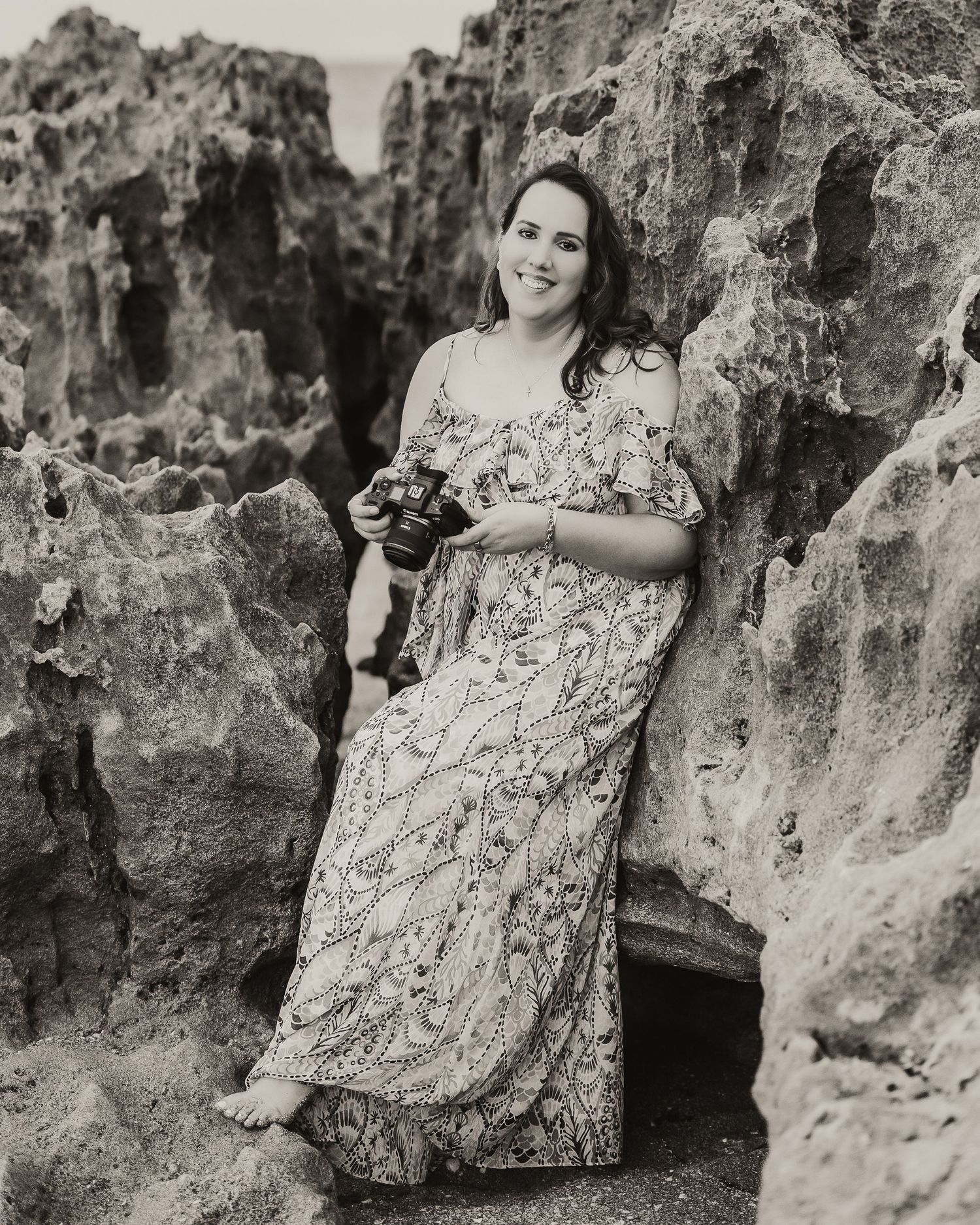A black and white portrait shows someone in a floral dress leaning against rocky formations outdoors.