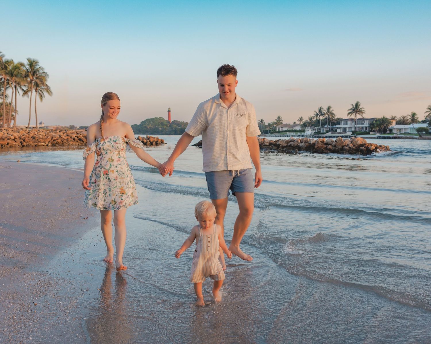 Three people walking hand in hand on a tropical beach at sunset with palm trees and boats in the background.