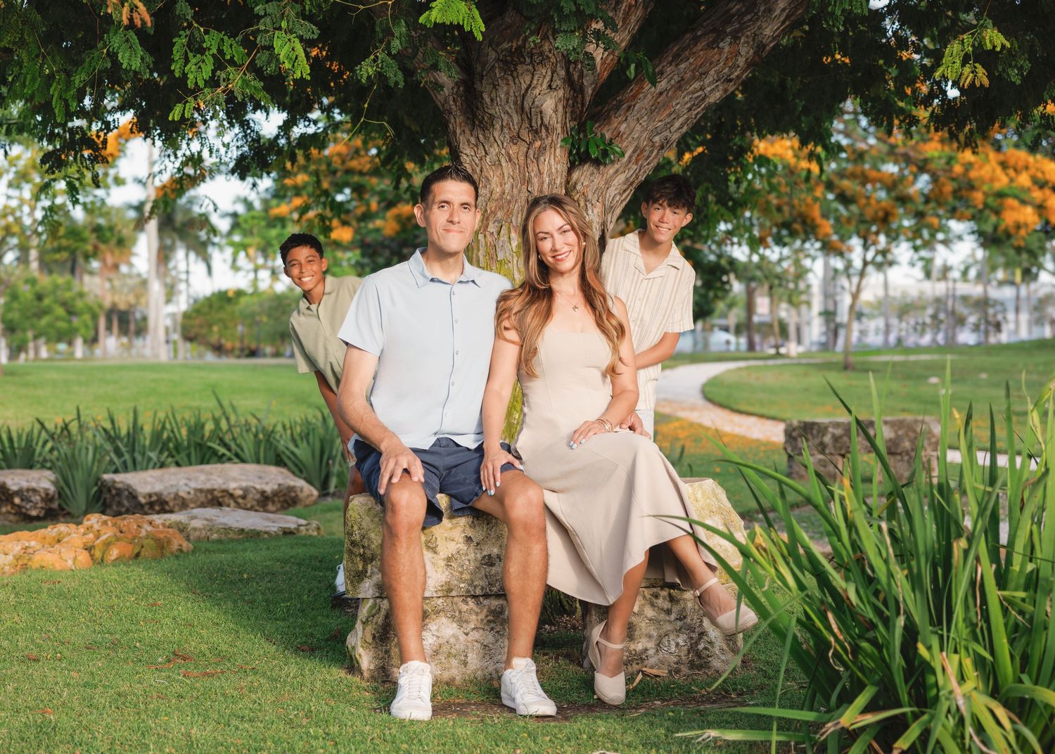 Family session sitting outdoors in a garden setting with lush greenery and a large tree during golden hour.