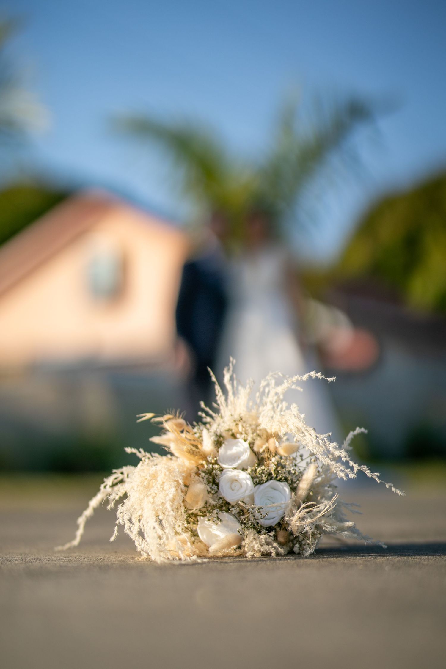 A dried flower wedding bouquet with pampas grass and white roses sitting on concrete in warm sunlight.