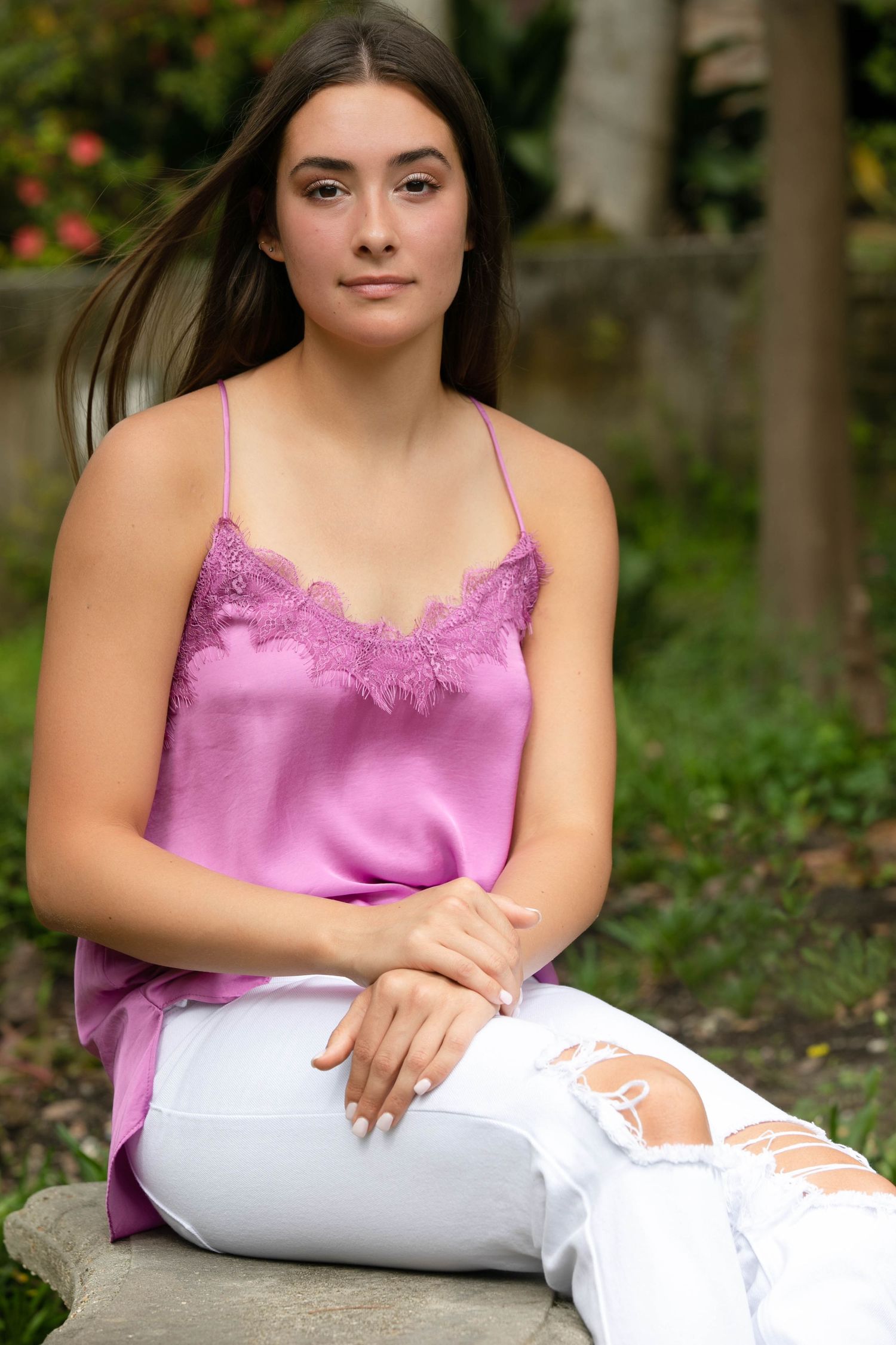 Portrait of someone in a pink lace camisole and white distressed jeans sitting outdoors against a natural background.