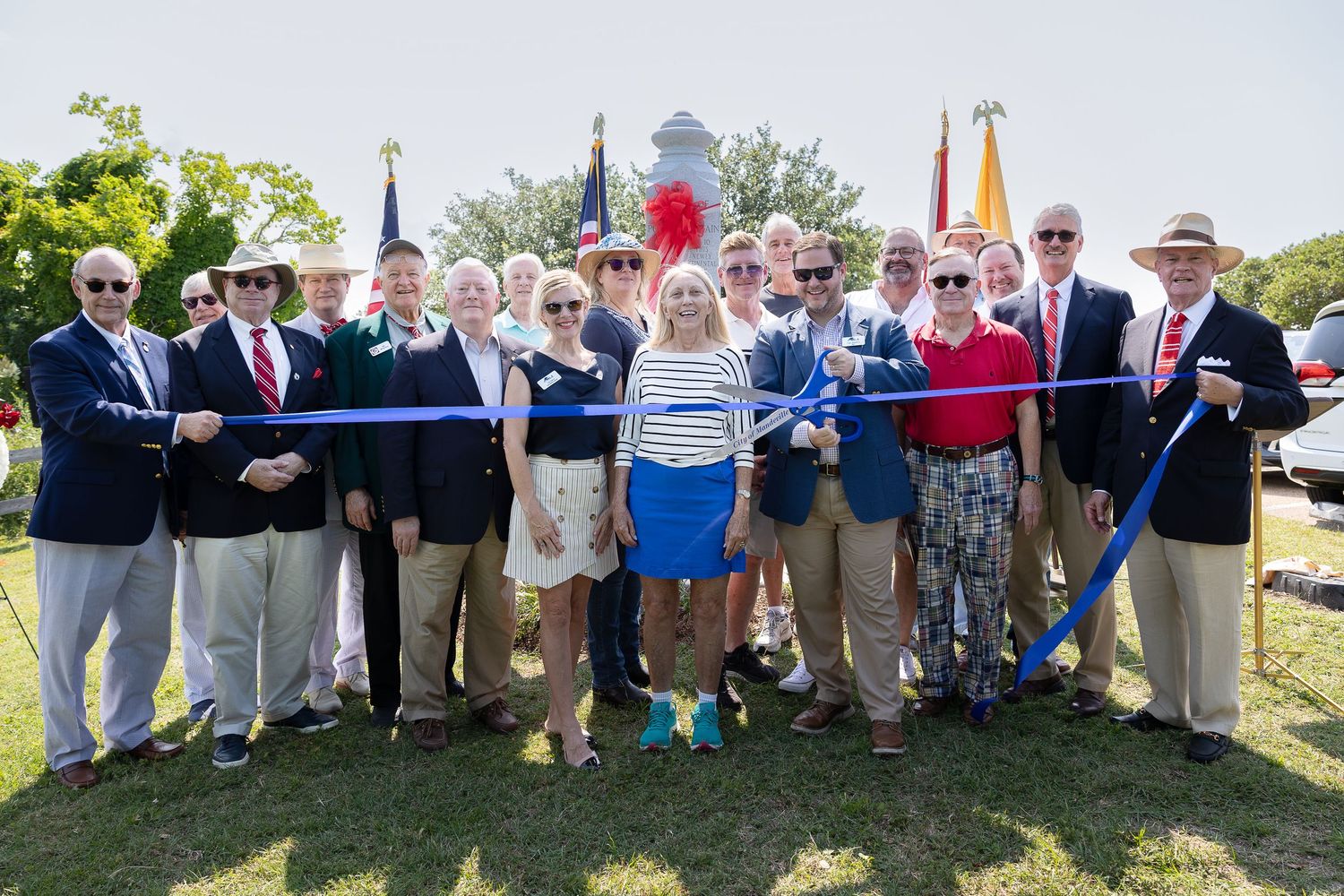 A group gathers outdoors holding a large ceremonial ribbon cutting for an event with red and blue ribbons. Mandeville Louisiana event photo