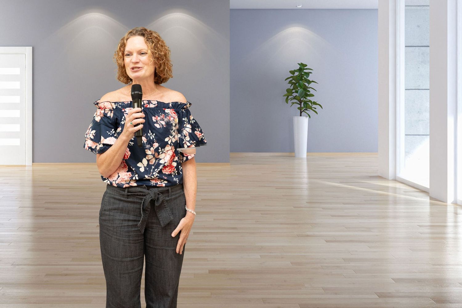 Someone in a floral off-shoulder top and dark pants speaks into a microphone in a bright modern space.