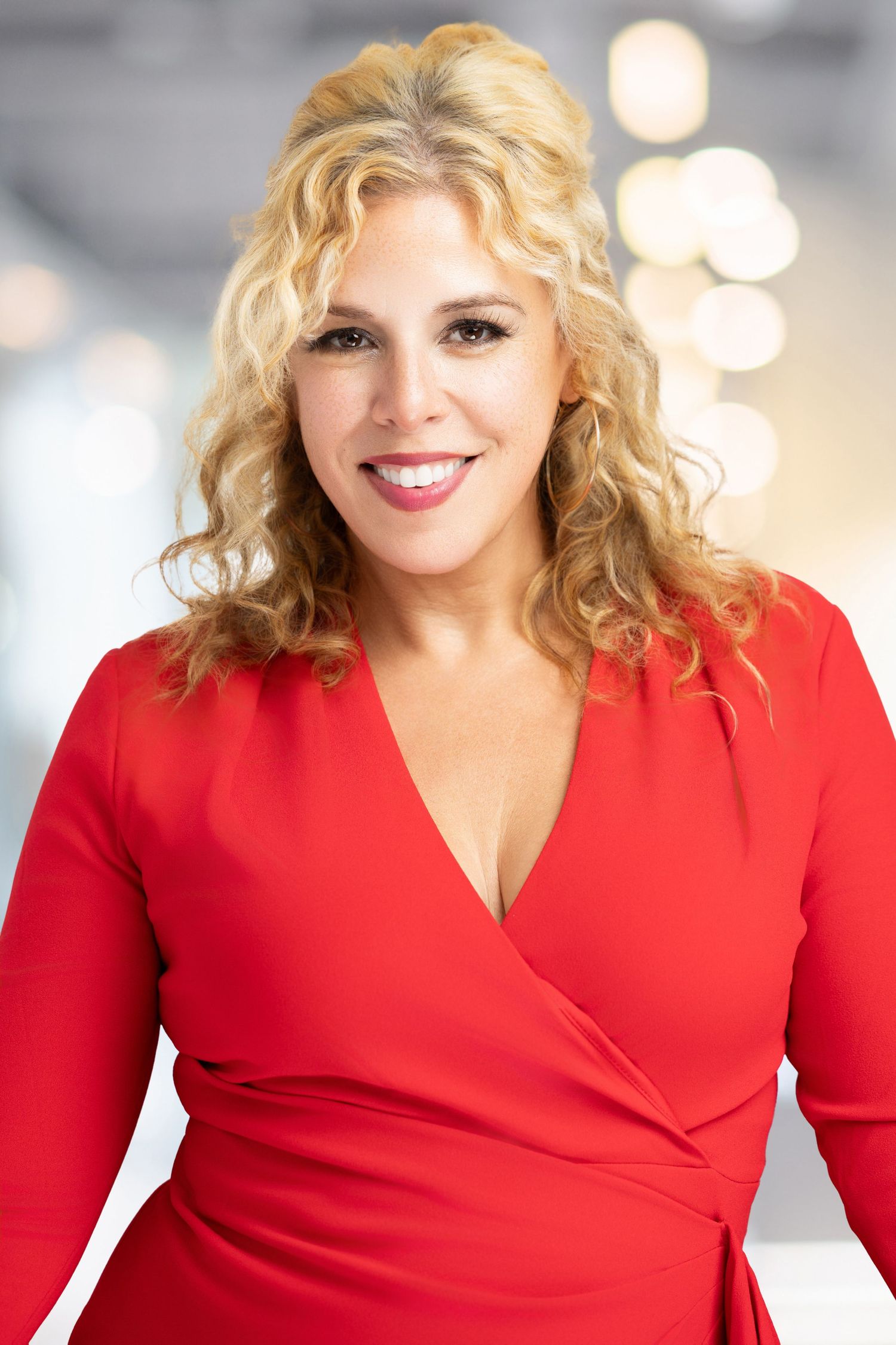 Professional portrait of a woman in a red wrap dress with curly blonde hair smiling against a blurred background.