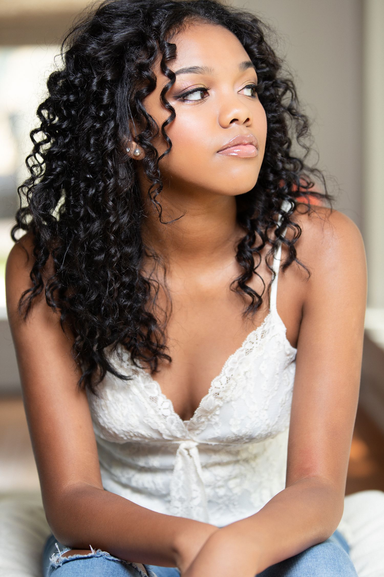 Elegant portraits showcase a woman in a white lace dress with natural curly hair posing in graceful poses near a window.