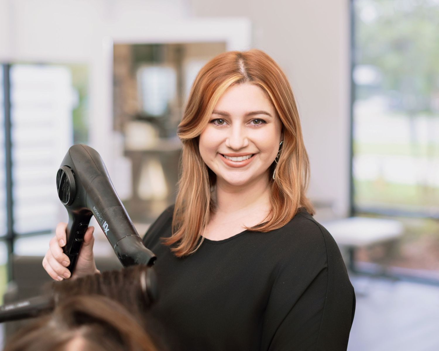 Professional hairstylist holding a blow dryer in a modern salon with bright natural lighting and blurred background. Louisiana branding.
