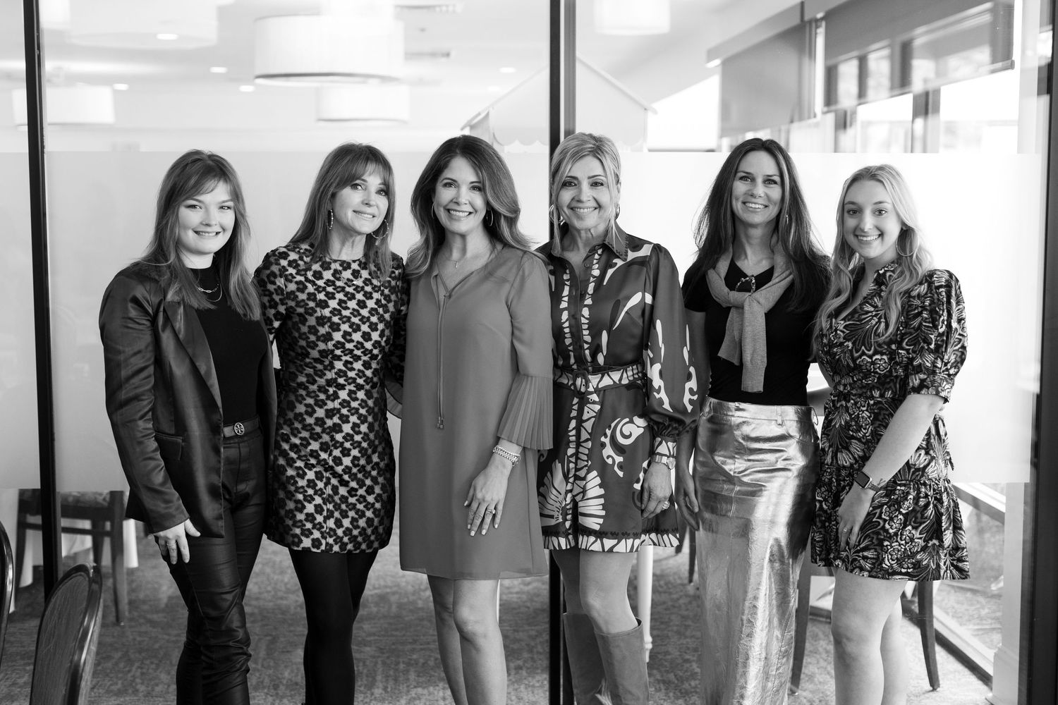 A black and white photograph shows a group of six friends posing together in a casual indoor setting.