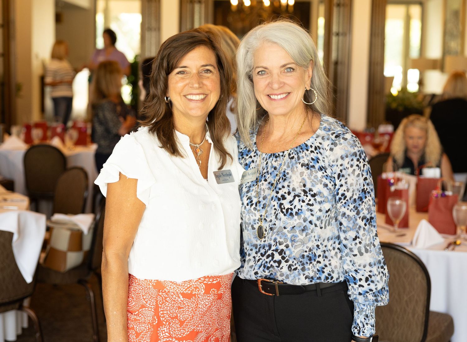Two women pose together at an elegant dining event, one wearing white top with coral skirt, another in floral print.